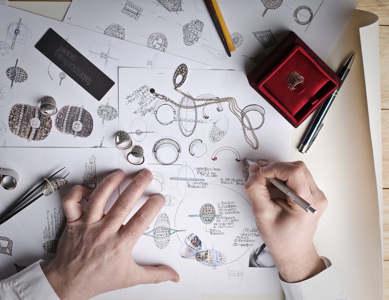 Hands sketching jewelry designs with tools and prototypes on a desk.